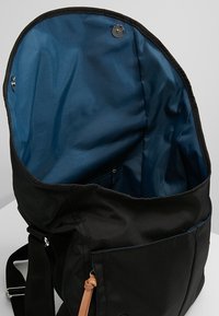 Borsa in tessuto nero con interni blu. Presenta una tasca con zip e un accento in pelle marrone sulla tracolla. Design con apertura superiore per un facile accesso.