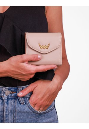 Compact beige wallet with a textured finish, gold zigzag emblem on the front, and a flap closure, held in a hand against denim.