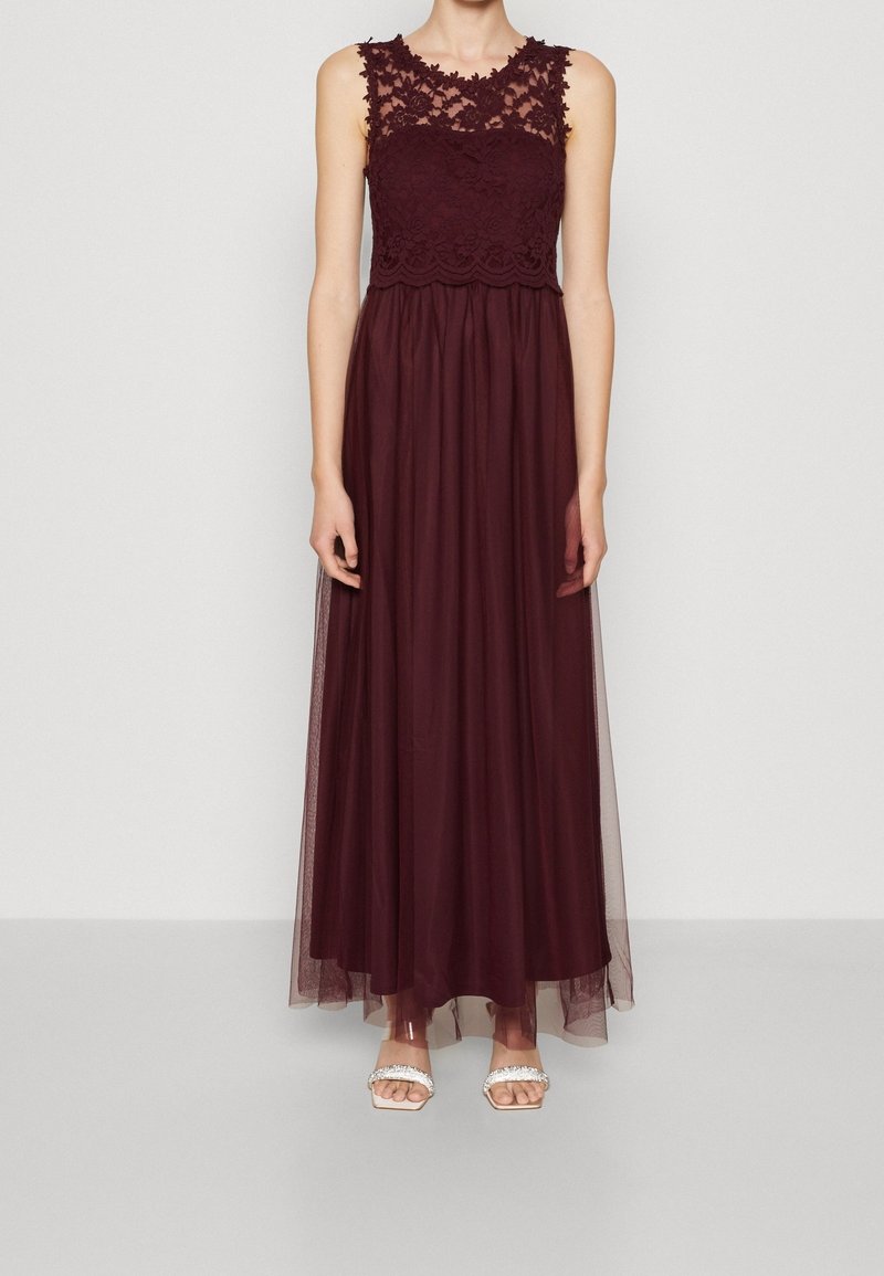 Robe de soirée bordeaux sans manches avec corsage en dentelle et superposition en tulle transparent, portée avec des escarpins blancs à bout ouvert.