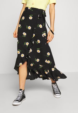 Jupe portefeuille noire avec imprimé marguerites blanches et ourlet à volants, portée avec des baskets montantes noires et un haut jaune à manches courtes.