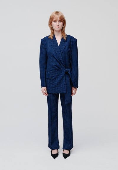 Donna con capelli rossi che indossa un blazer oversize a quadri blu con cintura e pantaloni abbinati, in piedi su uno sfondo grigio chiaro uniforme.