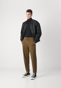 Schwarze Bomberjacke über einem schwarzen Rollkragenpullover, kombiniert mit braunen Jogginghosen mit Kordelzug und Logo. Getragen mit schwarz-weißen Turnschuhen.