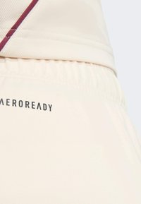 Zbliżenie beżowej elastycznej talii i części pasującej koszuli z bordowym paskiem oraz napisem "AEROREADY" na tkaninie.