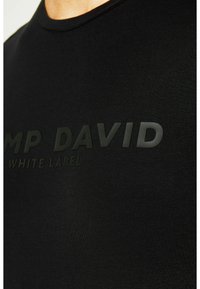 Camiseta negra de manga corta hecha de tela suave. Presenta un logo en relieve, en negro, de "CAMP DAVID WHITE LABEL" en la parte frontal. Diseño clásico de cuello redondo.