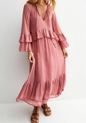 Kobieta ubrana w długą, lejącą się sukienkę w kolorze mauve z falbanami i rozszerzanymi rękawami, zestawioną z brązowymi, ćwiekowanymi płaskimi sandałami.