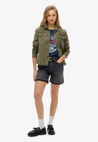 Chaqueta verde oliva con bolsillos frontales, camiseta gráfica azul, pantalones cortos de mezclilla negros con dobladillos deshilachados, calcetines blancos y zapatos negros.
