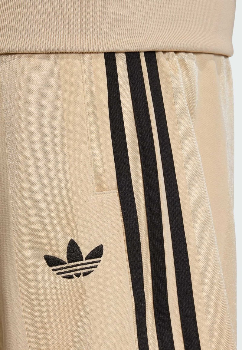 Pantalones deportivos beige con cintura acanalada, con franjas laterales negras y un logo negro de Adidas en el lado izquierdo. Textura suave.