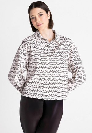 Jonge vrouw met kort donker haar, een wit overhemd met zwarte hartjespatronen en een zwarte broek, staand met handen achter de rug.