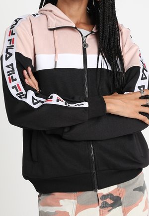 Osoba ubrana w czarno-różowo-białą bluzę z kapturem na zamek błyskawiczny z brandingiem FILA na rękawach, z rękami skrzyżowanymi, a w pasie widoczne spodnie moro.