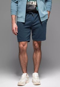 Męskie niebieskie spodenki z elastycznym pasem i sznurkiem, połączone z czarną koszulką z grafiką i beżowymi trampkami. Wykonane z miękkiej bawełny.