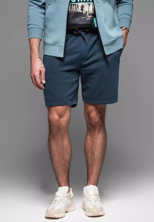 Shorts bleus pour hommes avec une taille élastique et un cordon de serrage, associés à un t-shirt à imprimé noir et des baskets beiges. Matériau en coton doux.