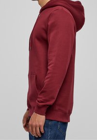 Sweat à capuche bordeaux avec une poche avant et une capuche à cordon, texture lisse, manches longues, et poignets et ourlet côtelés. Vue de côté présentée.