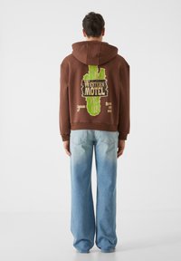 Ruda per didelė veliūrinė palaidinė su žalia grafika, kurioje užrašas "WESTERN MOTEL" ir kaktuso dizainas. Derinamas su šviesiai mėlynomis plačiomis džinsomis.