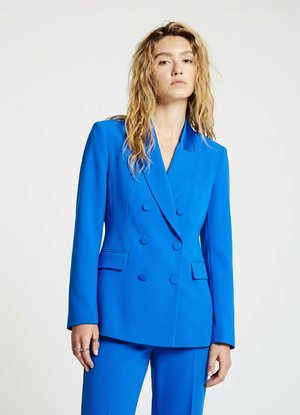 Donna con capelli biondi ondulati che indossa un blazer doppio petto blu brillante e pantaloni abbinati, in piedi davanti a uno sfondo semplice.