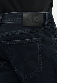 Μαύρο τζιν από denim με πίσω τσέπη και μαύρη δερμάτινη ταμπέλα της Calvin Klein στη μέση της μέσης, φορεμένο με μαύρο τοπ.