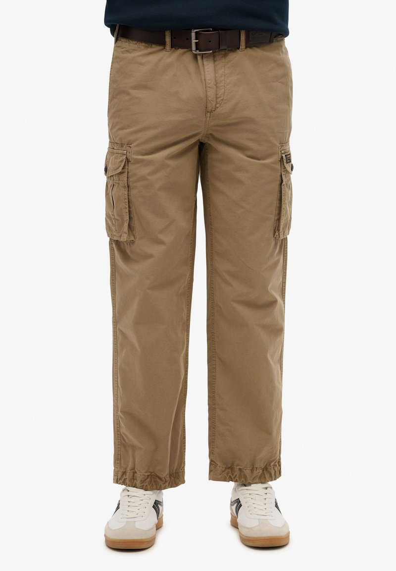 Pantaloni cargo beige con tasche laterali, vestibilità dritta, polsini elasticizzati e tessuto in cotone; indossati con sneaker bianche con accenti neri.