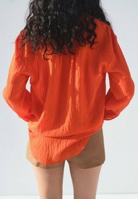 Orange krinklad långärmad skjorta med lös passform, med en rundad nederkant och en strukturerad tyg, matchad med beiga shorts.