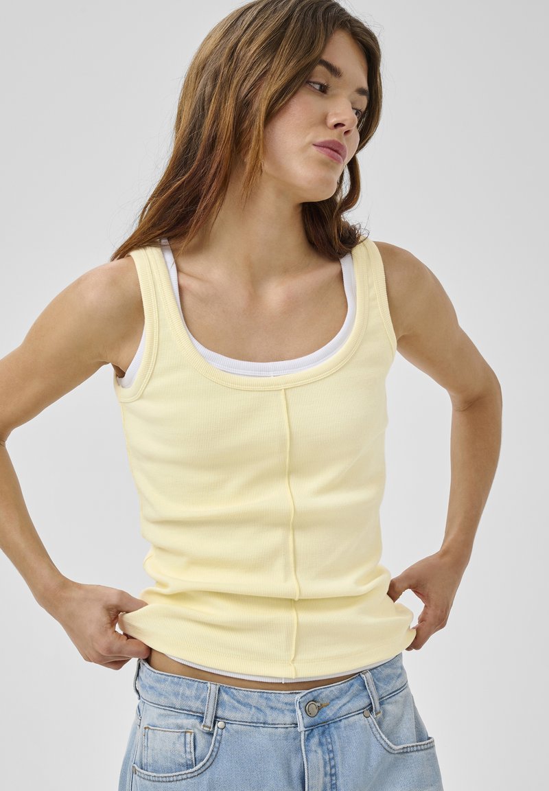 Jeune femme ajustant un haut jaune sans manches par-dessus un sous-vêtement blanc, portant un jean bleu clair, regardant sur le côté sur un fond neutre.