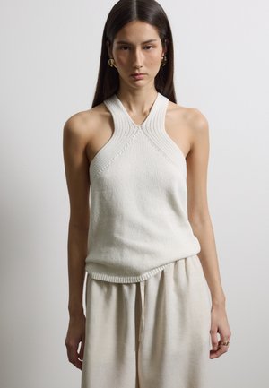 Frau trägt ärmelloses, leichtes Strickoberteil mit Neckholder und lockere beige Hose, steht vor schlichtem hellgrauem Hintergrund.