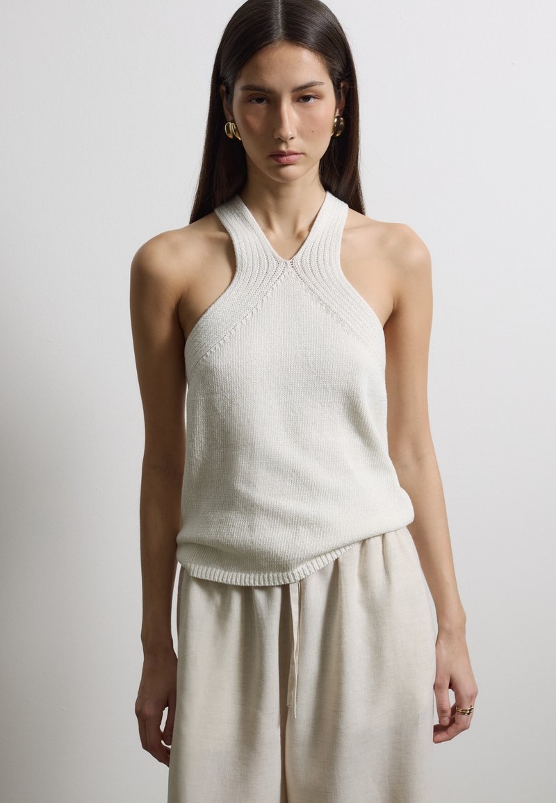 Frau trägt ärmelloses, leichtes Strickoberteil mit Neckholder und lockere beige Hose, steht vor schlichtem hellgrauem Hintergrund.