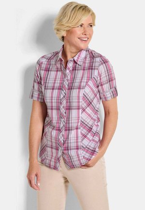 Femme aux cheveux blonds courts portant une chemise à manches courtes à carreaux rose et blanc et un pantalon beige, souriante et regardant sur le côté.