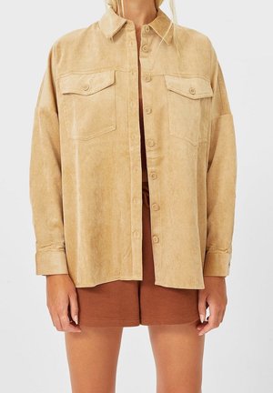 Femme portant une chemise boutonnée en velours côtelé beige oversized par-dessus un short marron, les mains détendues le long du corps sur fond blanc.