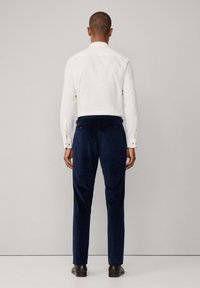 Bijela košulja s dugim rukavima u kombinaciji s tamnoplave kord flaširane hlače, s ravnim nogavicama, bočnim džepovima i tamnosmešnim cipelama.