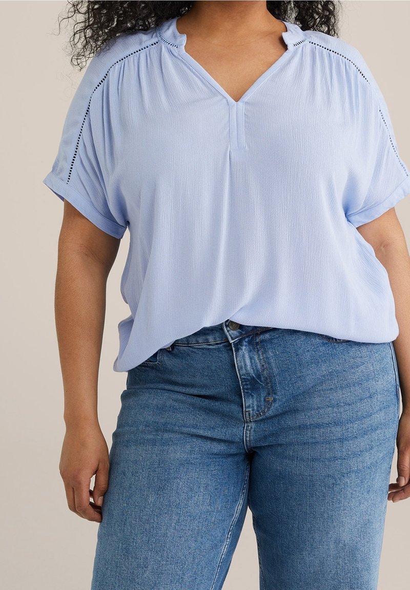 Femme portant un chemisier bleu clair à manches courtes avec col en V et détails en dentelle, associé à un jean en denim bleu taille haute.
