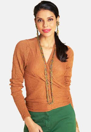 Frau mit dunklen Haaren, braunem Langarmshirt und grüner Hose, die eine lange bunte Perlenkette und passende Blumenohrringe trägt, lächelt sanft.