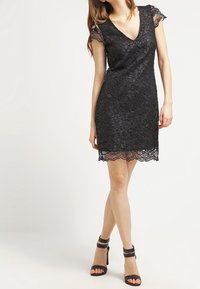 En kvinna iklädd en svart spetskortärmad V-ringad klänning och svarta högklackade sandaler, stående med benen korsade mot en vit bakgrund.