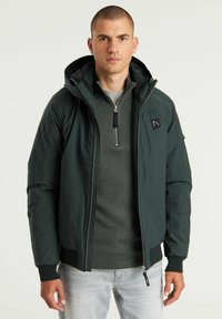 Chaqueta aislante de color verde oscuro con capucha, cierre de cremallera y puños acanalados. Presenta un bolsillo lateral y un parche de logo en el pecho.