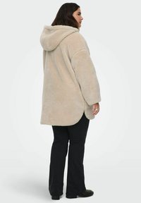 Bežs flīsa pārmērīgs apģērbs ar kapuci, ar mīkstu tekstūru, garām piedurknēm un noapaļotu apakšējo malu. Saskaņots ar melnām biksēm un apaviem.