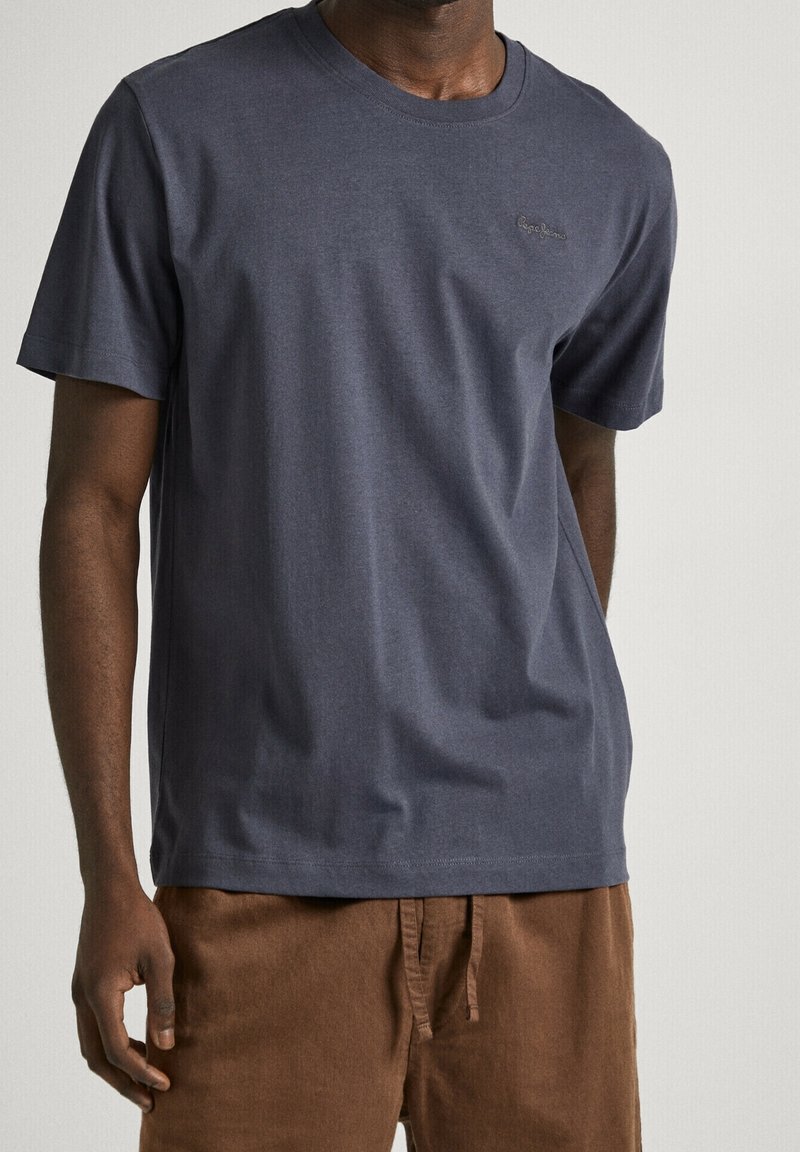 Homme portant un t-shirt gris foncé uni à manches courtes et un pantalon marron sur un fond clair neutre.