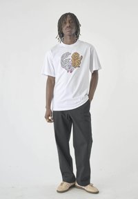 Mann mit Dreadlocks, der ein weißes T-Shirt mit einem Cartoon-Katze und -Hund trägt, schwarze Hose, beigefarbene Sneakers, steht mit einer Hand in der Tasche, weißer Hintergrund.