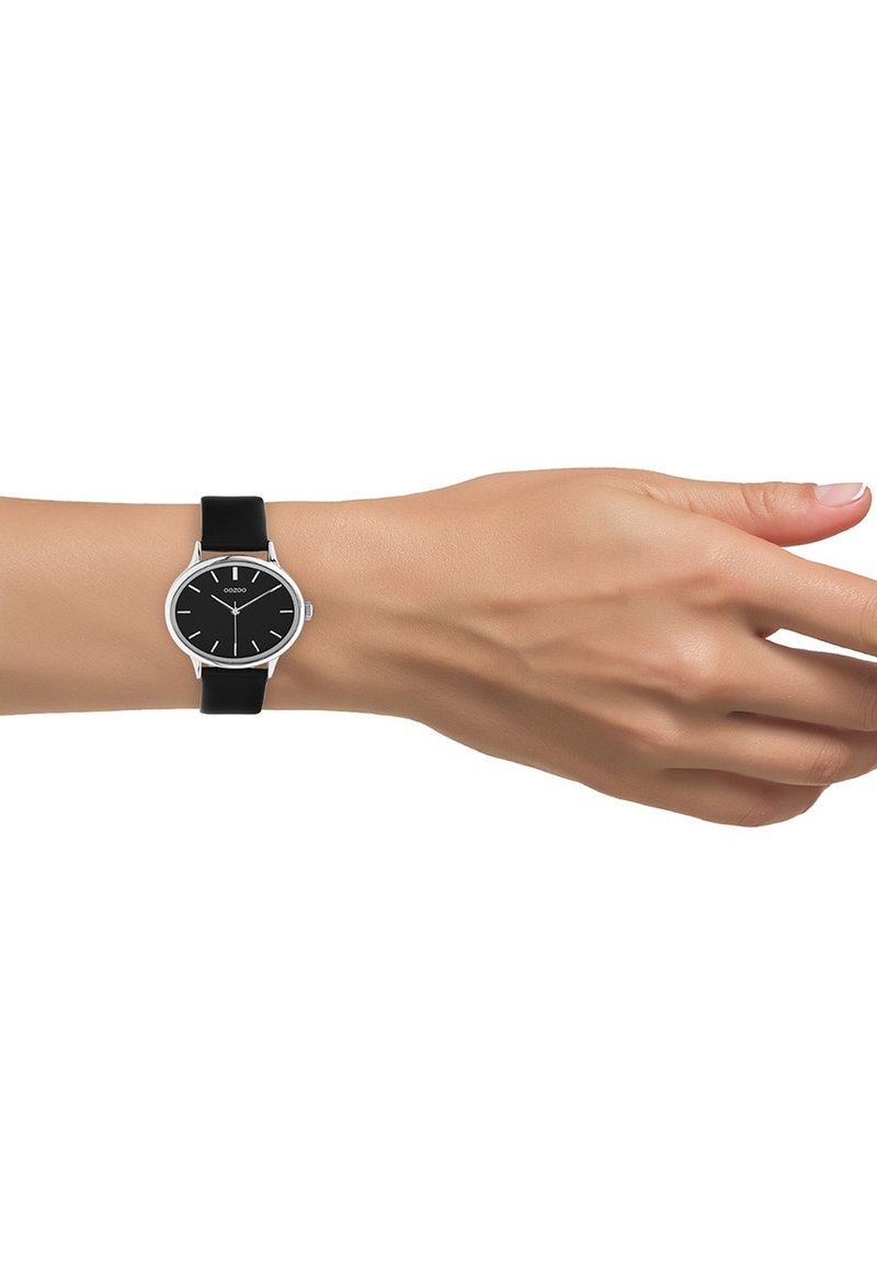 Montre noire avec boîtier en acier inoxydable rond, cadran noir, index argentés et bracelet en silicone noir, présentée sur une main.