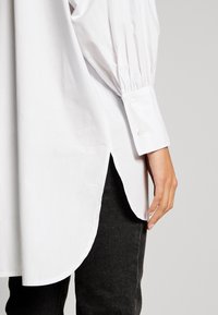 Camicia bianca a maniche lunghe con design a maniche plissettate, orlo arrotondato e polsini abbottonati, indossata sopra dei jeans scuri. Tessuto morbido e leggero.
