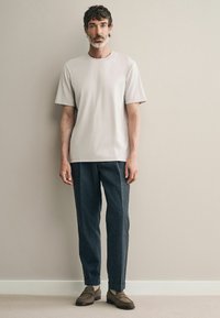 Jasnoszara koszulka z krótkim rękawem, ciemne teksturowane spodnie oraz brązowe buty typu slip-on, na jednolitym tle w neutralnych tonach.