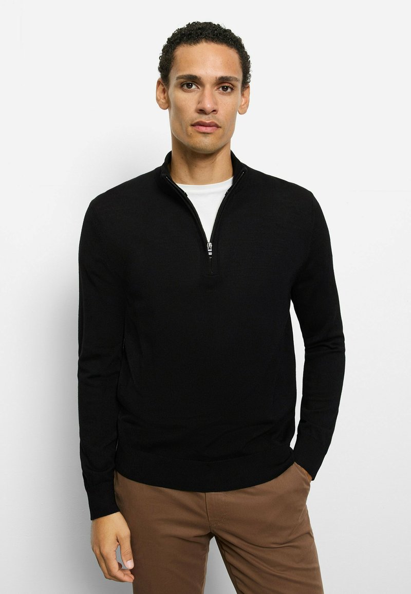 Schwarzer Pullover mit Viertelreißverschluss am Kragen, gerippten Bündchen und Saum, gefertigt aus einem strukturierten Stoff. Über einem weißen T-Shirt getragen.