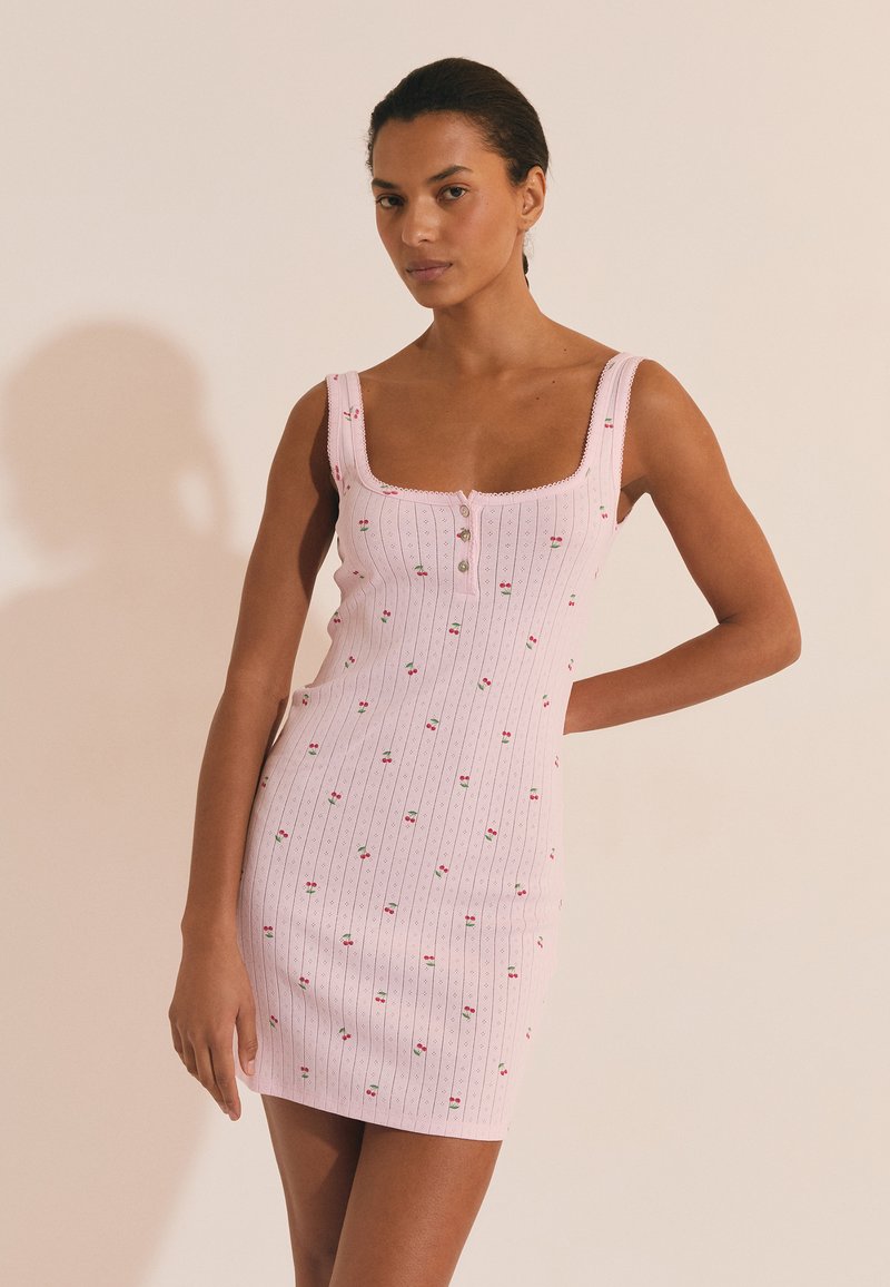 Femme portant une mini-robe rose sans manches avec de petits motifs de cerises et des boutons sur un fond beige clair.