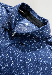 Tummansininen lyhythihainen paita, joka on valmistettu teksturoidusta kankaasta. Paidassa on toistuva kuvio vaaleammissa sinisissä sävyissä ja vetoketjukaulus.