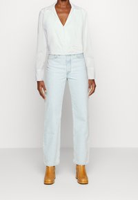 Abgeschnittenes weißes Hemd mit V-Ausschnitt und langen Ärmeln, kombiniert mit hellblauen, hochgeschnittenen Jeans mit geradem Bein und beigefarbenen Stiefeletten mit Absatz.