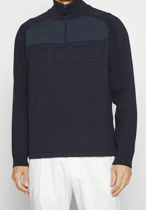 Mand iført en marineblå pullover med kvartlynlås og præget "BOSS" logo over brystet, kombineret med hvide bukser.