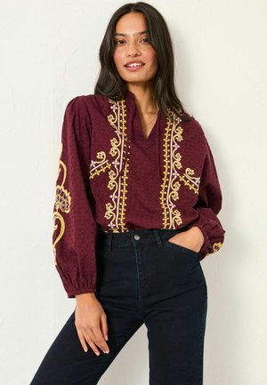 Burgundy textured blouse with yellow and white floral embroidery, featuring a v-neck and voluminous sleeves. Paired with dark jeans.