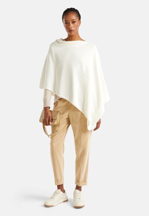 Un poncho en laine blanche drape sur un pull beige, associé à un pantalon en velours côtelé clair et des baskets crémeuses. Accessoirisé avec un sac à main en chaîne dorée.