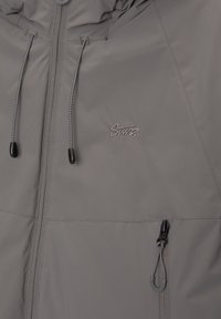 Veste grise zippée avec cordons de capuche réglables, fermeture éclair avant, poche latérale zippée et petit logo brodé sur la poitrine.