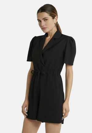 Frau mit heller Haut und braunen Haaren in niedrigem Pferdeschwanz trägt schwarzen, kurzärmligen, taillierten Overall mit Reverskragen, steht vor weißem Hintergrund.