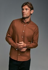 Camisa marrón de botones con cuello, que presenta botones blancos y un pequeño logotipo bordado en el pecho, combinada con pantalones oscuros.