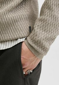 Pull en tricot marron clair avec un motif texturé, associé à une chemise blanche à rayures, un pantalon foncé et des bagues en argent sur une main.