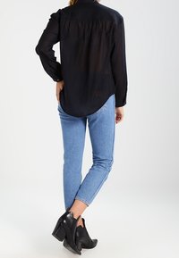 Black, long-sleeve, sheer shirt with back pleats, paired with blue jeans and black heeled ankle boots. Simple design, no patterns visible.