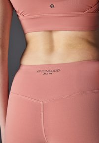 Svetloružový športový set obsahujúci priliehavú športovú podprsenku a legíny s vysokým pásom, vyrobené z hladkého materiálu a s minimalistickým logom na páse.